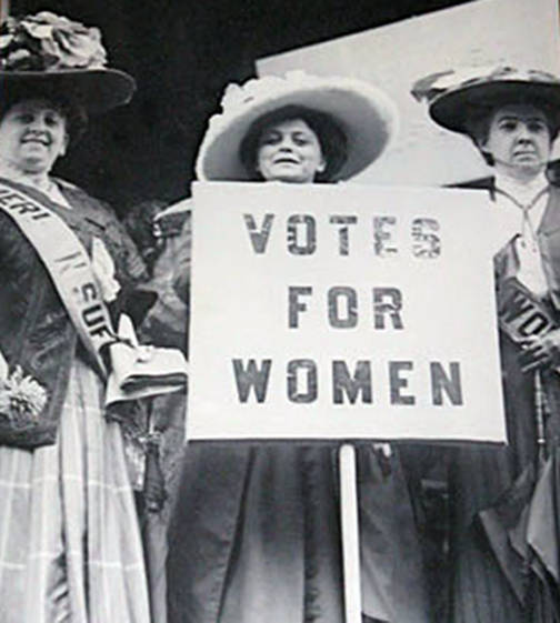 Three suffragettes hold up a sign that says, 'votes for women'